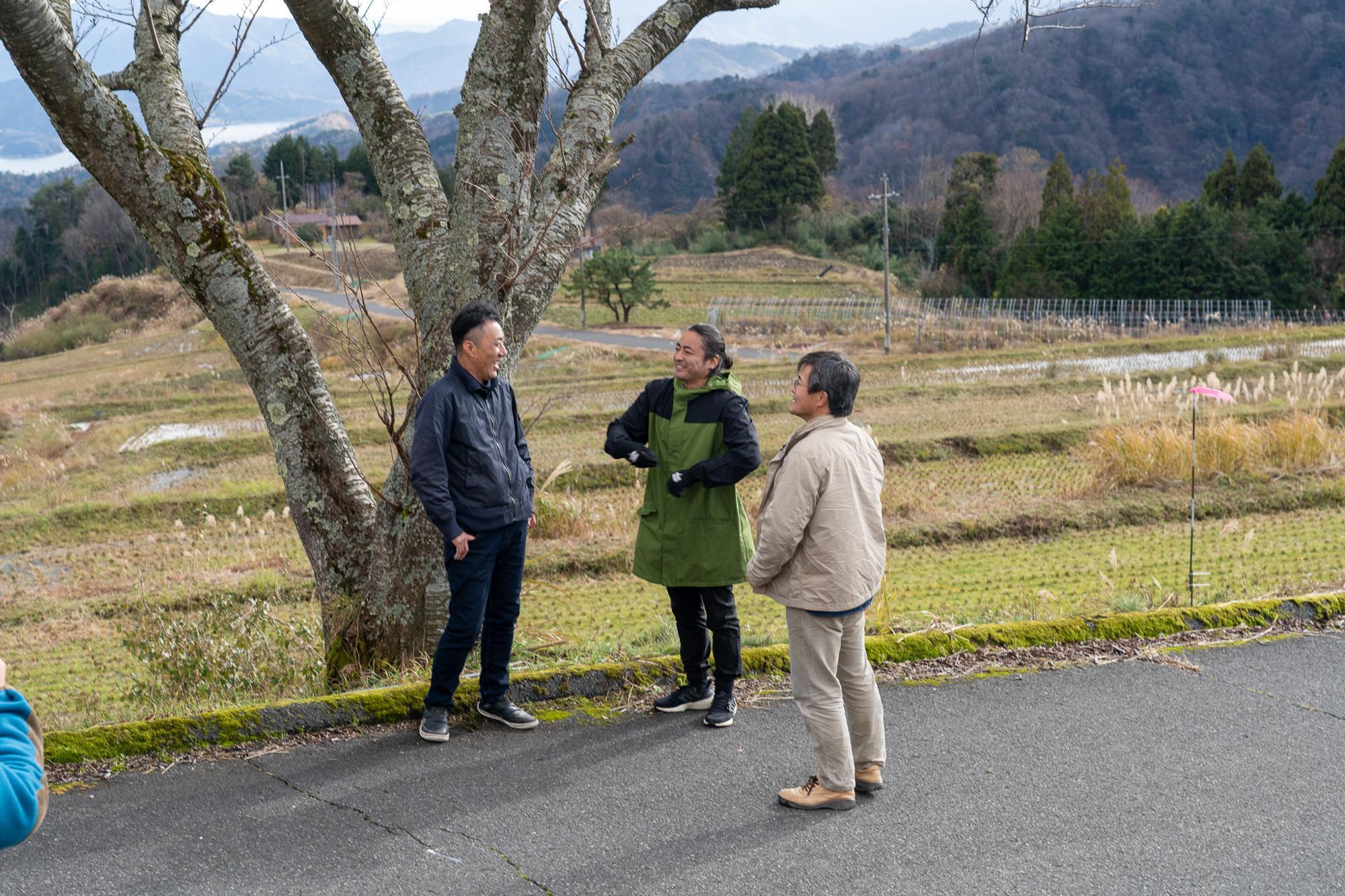 『原点回帰』で稲作を始める田んぼの前で。目印となる大きな一本桜の木の下で飯尾醸造で米づくりを担当する伊藤浩二さんと農家の溝口嘉順さんと談笑