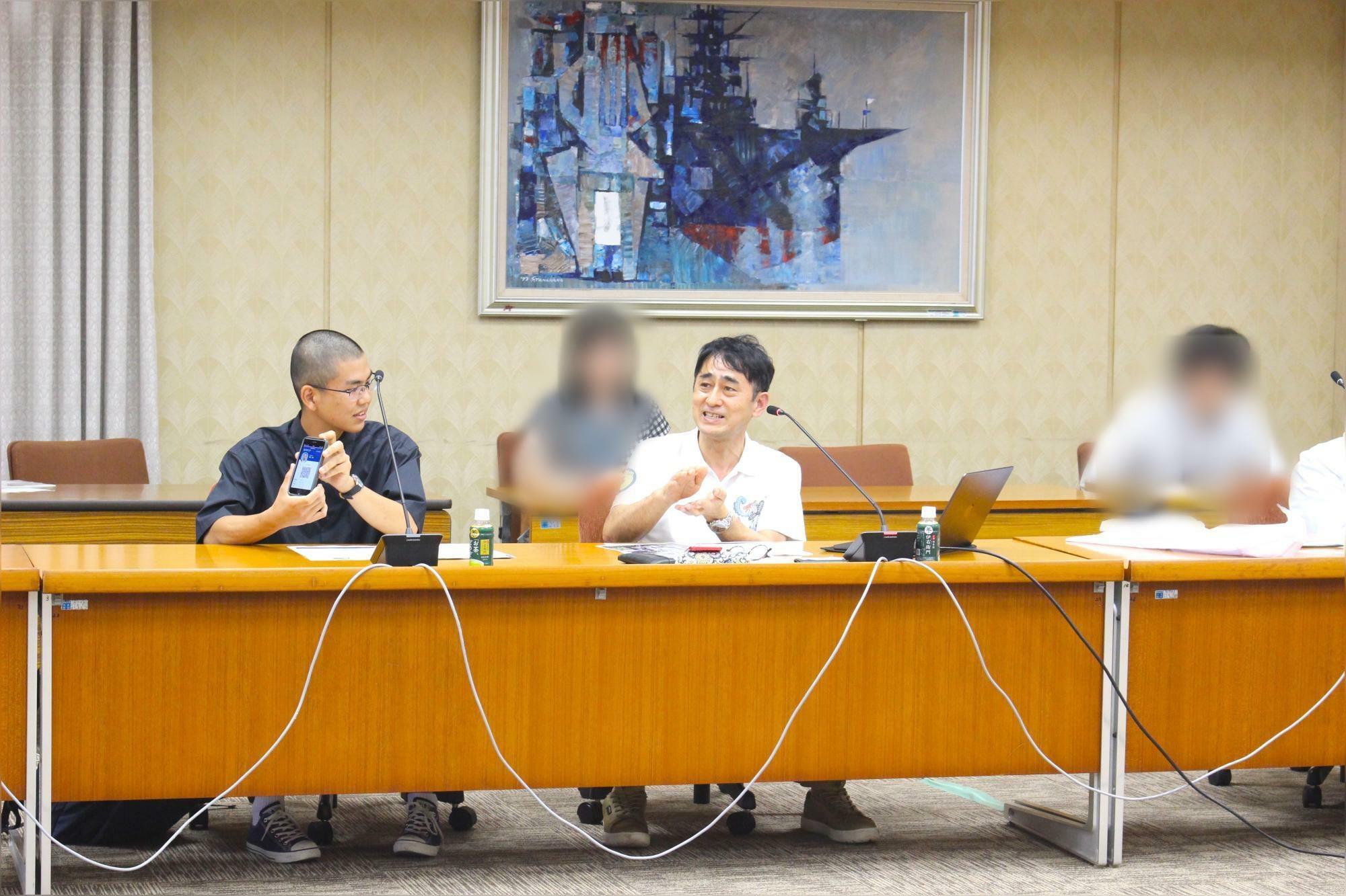 画面中央：記者会見にてアプリの説明を行う岡﨑威生 教授（琉球大学 工学部）