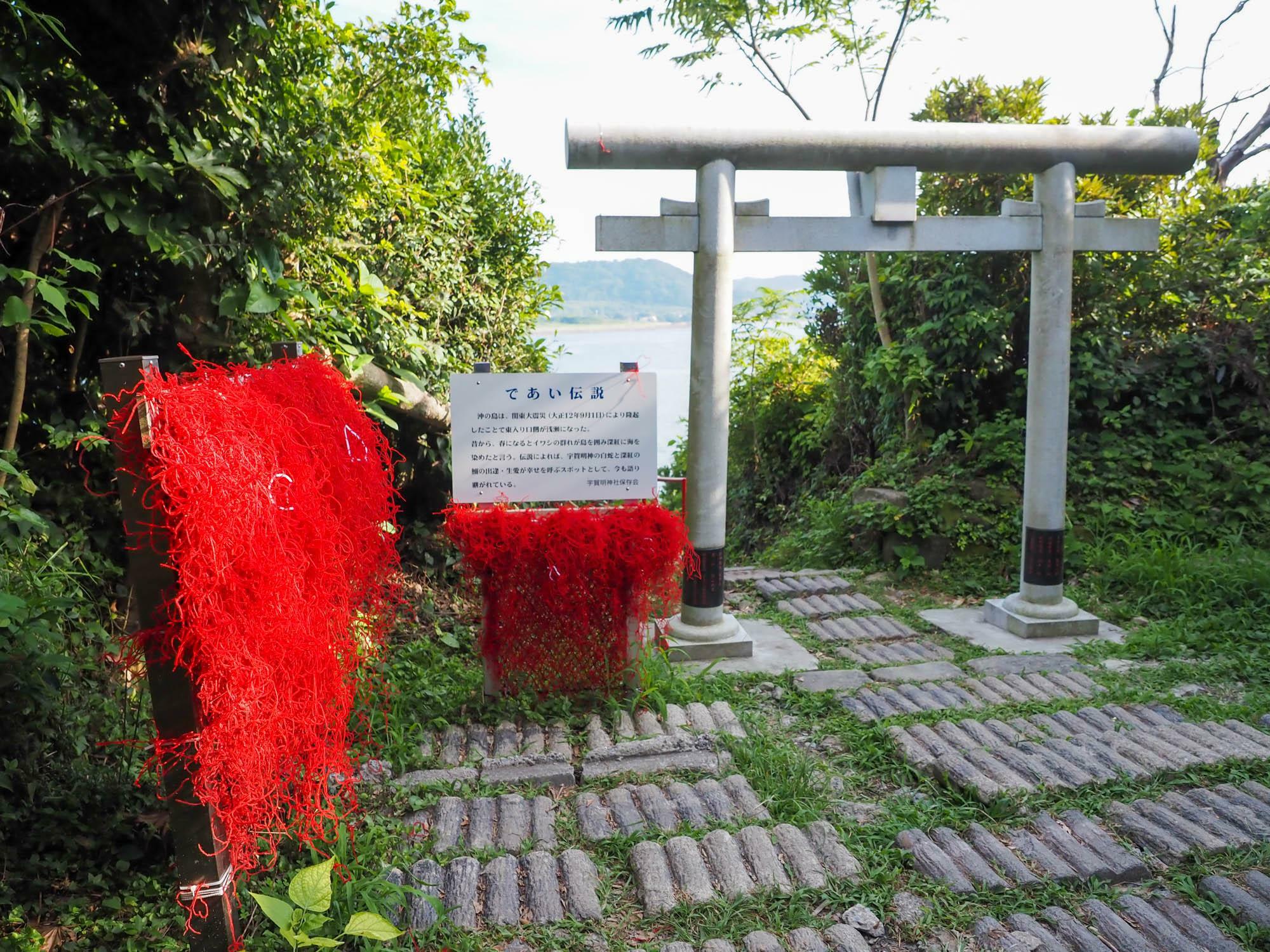 沖ノ島宇賀大明神社。真っ白の鳥居越しに青い海が見える。その鳥居の側に設置された「いわし網」にはたくさんの赤い糸が結び付けられていて、白と赤の色の対比が鮮烈である。