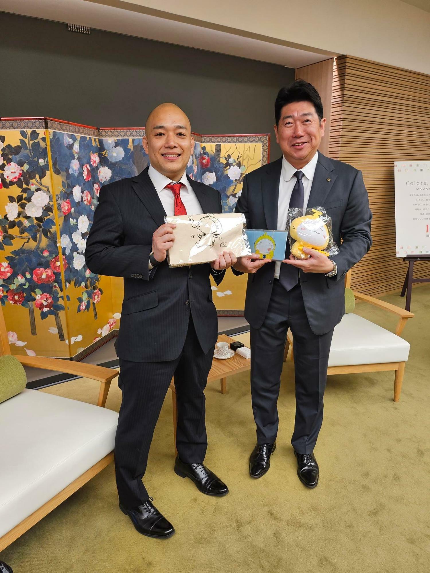 オズ通り商店街のマスコットキャラクター・おずっちょグッズを持つ岩佐理事長（左）と福田市長