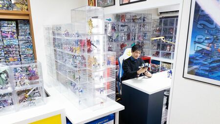 タカハシさんがコレクションに囲まれた趣味部屋に立つ様子。膨大なガンプラやグッズが壁一面に並ぶ、理想のガンダム空間。