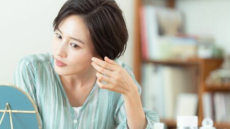 白髪の根元が伸びてきて、女性が不安そうな表情で自分の髪を見ている様子。頻繁な白髪染めの必要性と「白髪に追われる」感覚を示唆している。