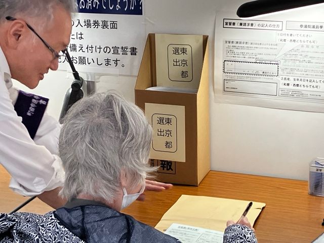 選挙管理委員会のスタッフに付き添われ、投票用紙を受け取る高齢の女性。車椅子で投票所に到着した様子を示しており、身体の不自由な方への支援の重要性を強調しています。