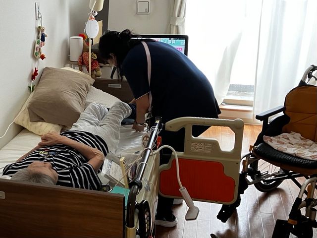 都内の施設に入居する要介護4認定の母親がベッドに横たわり、身体的な制約から日常動作にも介助が必要な状態を表しています。郵便投票制度の対象外となる高齢者の困難を示唆。