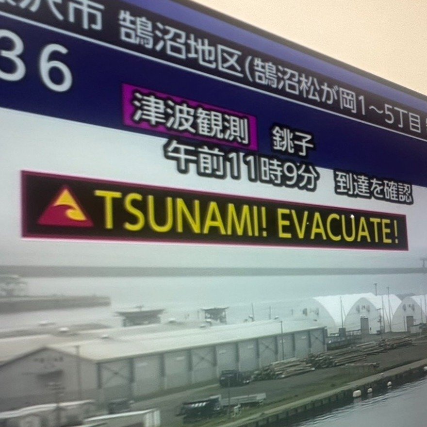NHKの津波警報テロップ、英語表記「TSUNAMI」「EVACUATE」が交互に表示されているテレビ画面。