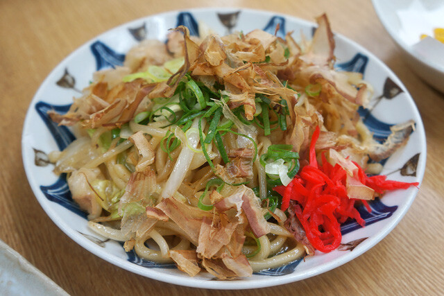 大阪・千林の老舗酒場「春倉」で提供される名物「ホルモン焼うどん」の写真。食欲をそそる香ばしいホルモンとうどんの組み合わせが、活気ある店内を背景に写し出されている。