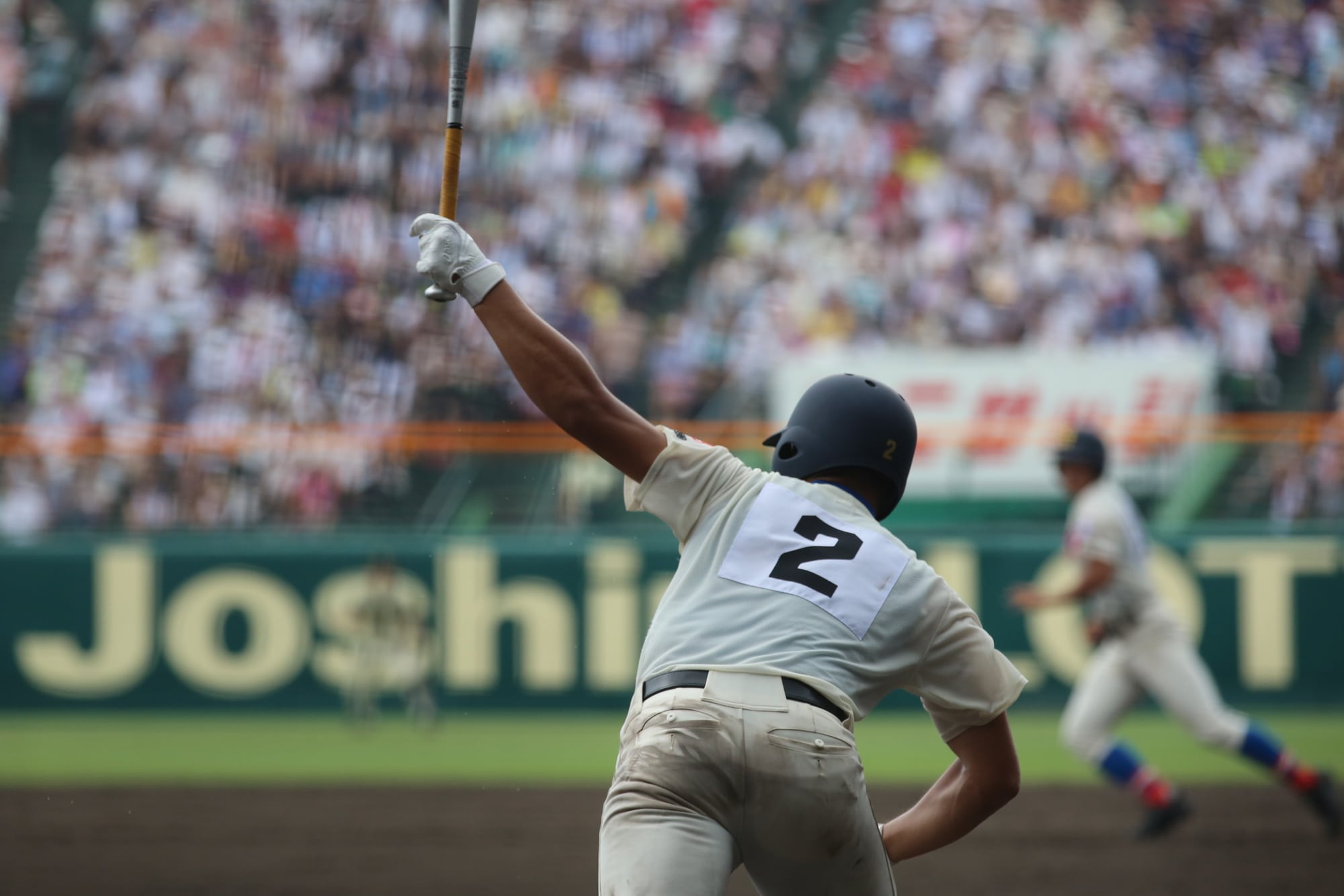 青空の下、野球場で練習する高校球児たちの後ろ姿。甲子園の華やかな舞台の裏側にある「現実」を象徴する。