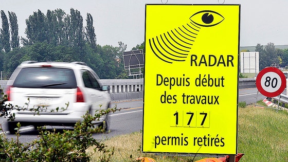 スイスの高速道路脇に設置された、スピード違反による運転免許取消件数を示す警察の表示板と通過する車。スイスの厳格な交通法規と罰金制度を象徴。