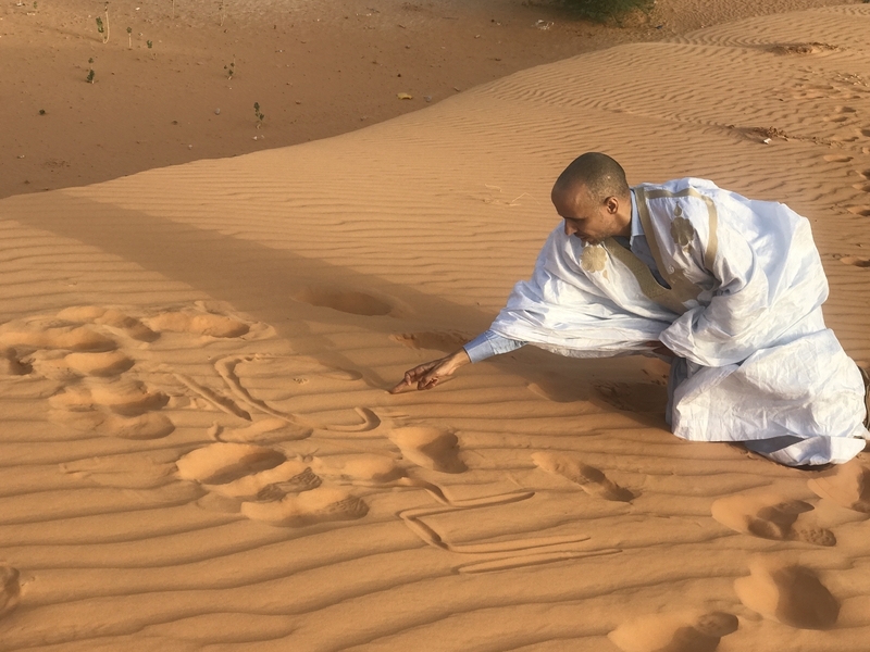 モーリタニア・ヌアクショット郊外の砂漠で、「神の御加護を」とアラビア語で砂に書くモハメドゥ（筆者撮影、２０１７年）