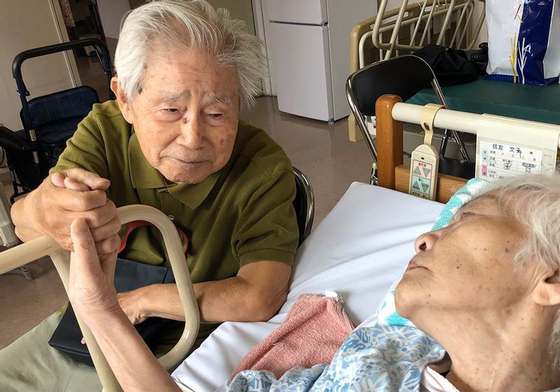 入院先を見舞い、妻の手を握って語りかける信友良則さん=(C)2022「ぼけますから、よろしくお願いします。~おかえり お母さん~」製作委員会