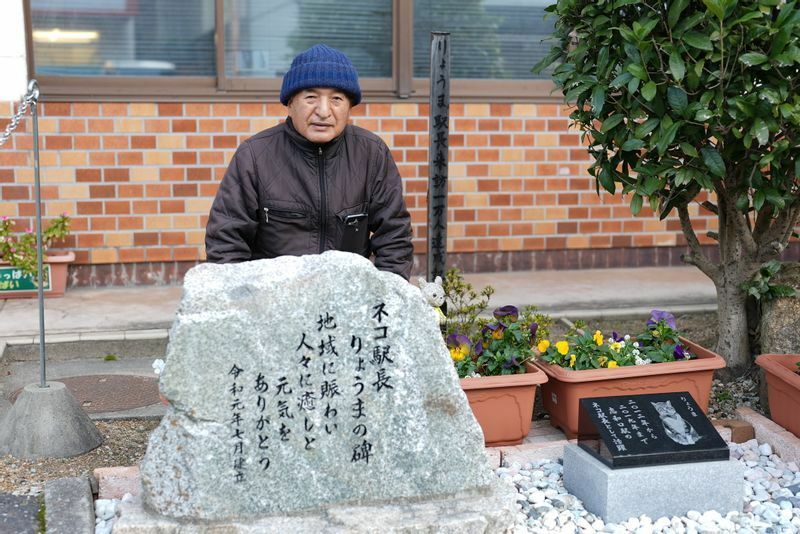 志和口駅前にある「りょうまの碑」と中原さん。「りょうまの死後、駅を訪れたファンから『何か功績を伝えるものを』との声が相次ぎ、地元産の花こう岩に謝辞を刻んで建立しました」（中原さん）