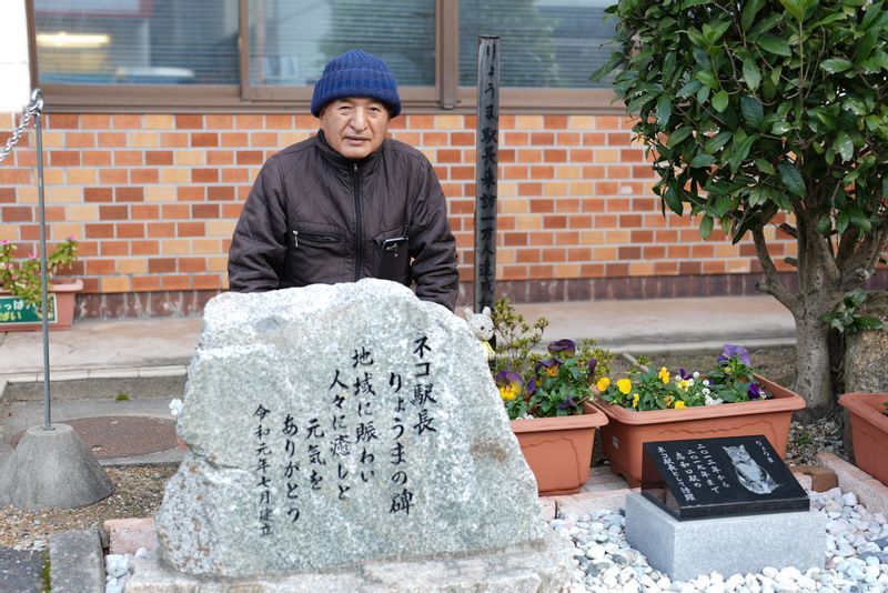 志和口駅前にある「りょうまの碑」と中原さん。「りょうまの死後、駅を訪れたファンから『何か功績を伝えるものを』との声が相次ぎ、地元産の花こう岩に謝辞を刻んで建立しました」(中原さん)