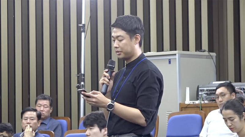 2日の記者懇談会で、携帯電話片手に質問する韓国紙記者。与党・共に民主党のYouTube中継よりキャプチャ。