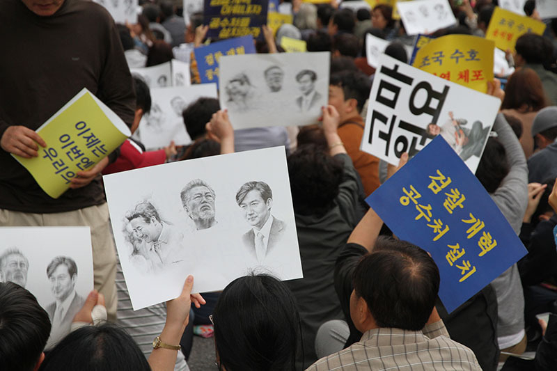 5日の進歩派デモで最も多く見かけたプラカード。左から故盧武鉉大統領、文大統領、チョ氏の顔が並ぶ。検察の侮辱的な捜査を苦に自殺した盧元大統領を連想させ、右の二人を守り抜くという強い意志表示だ。筆者撮影。