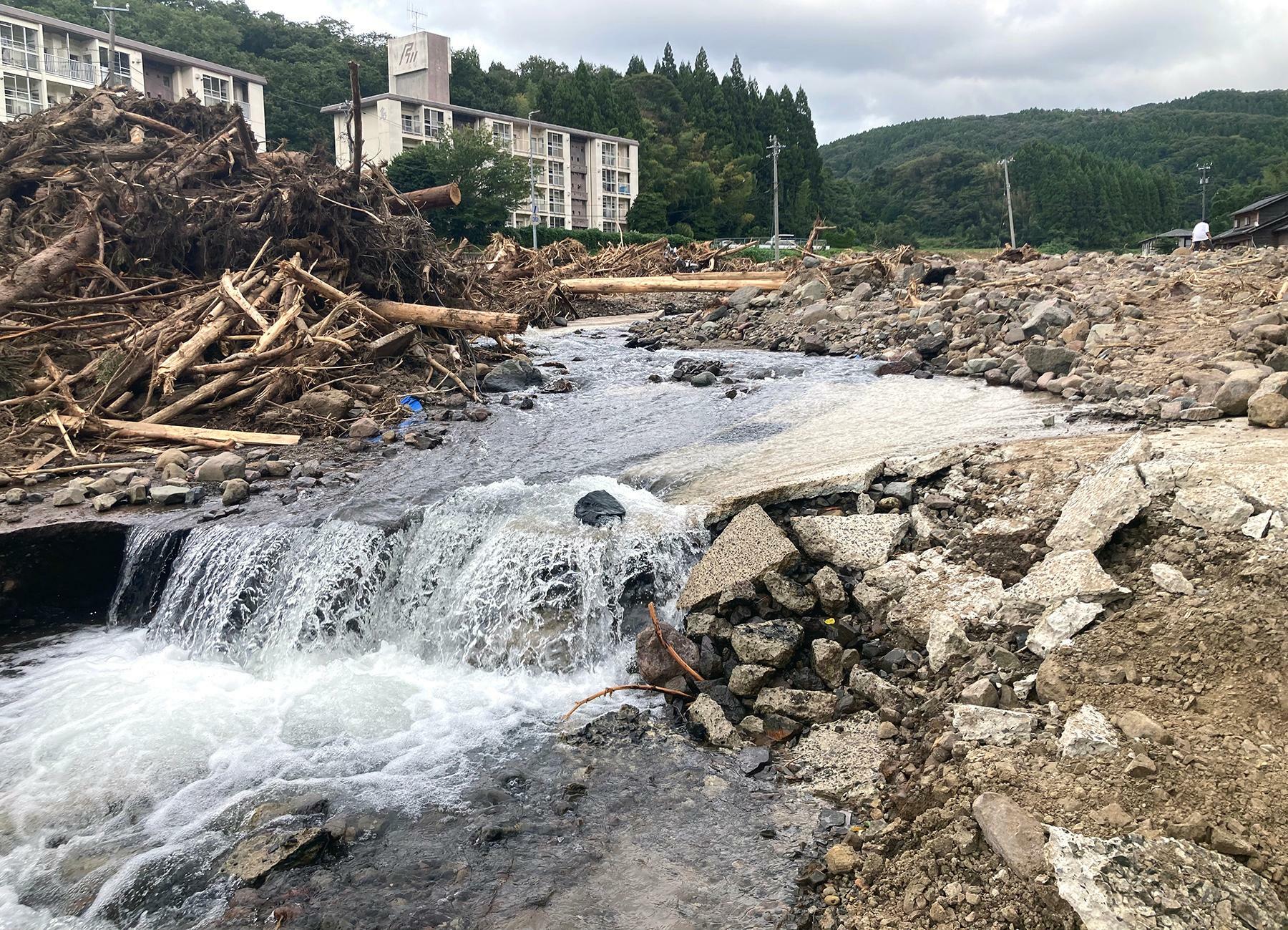 輪島市久手川町の被災現場。水が流れているのは元の道路。奥には市営・県営住宅が並ぶ久手川団地が見える＝9月28日、筆者撮影