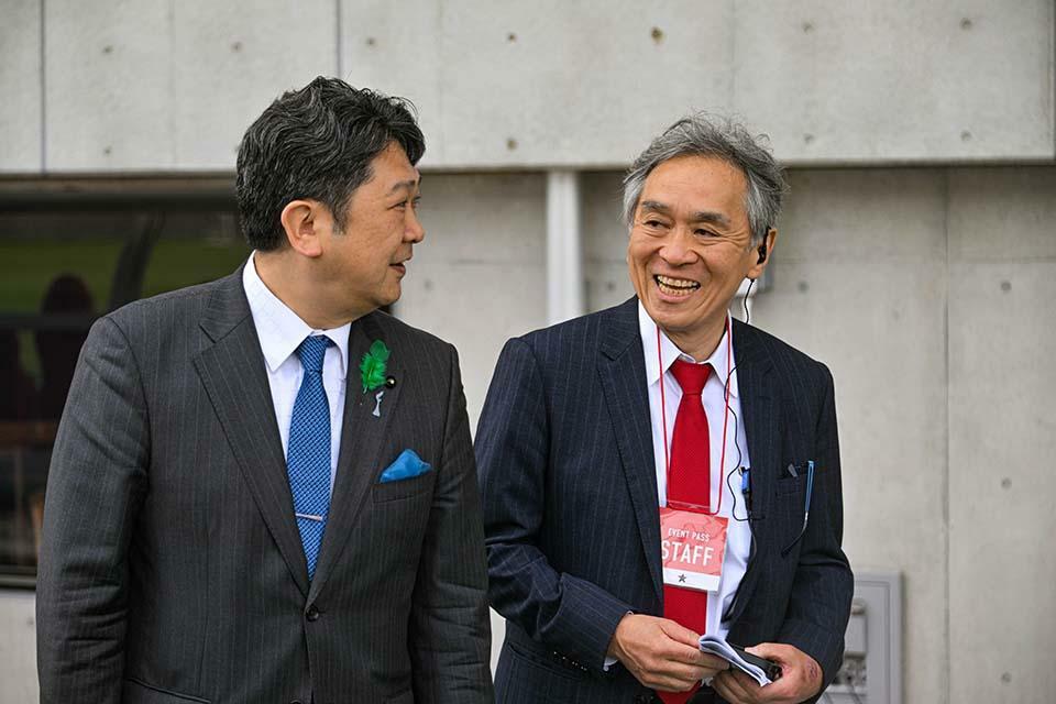 金沢市の村山卓市長と談笑する小島潔氏（右）。今回のチャリティーマッチは、この人物の強い想いが起点となっている。