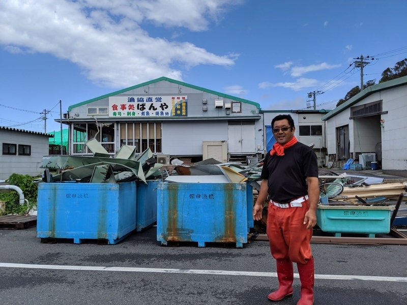 保田漁協の鈴木さん。後ろに積まれているのはばんやの建物の屋根だったがれき（筆者撮影。2019.10.4）